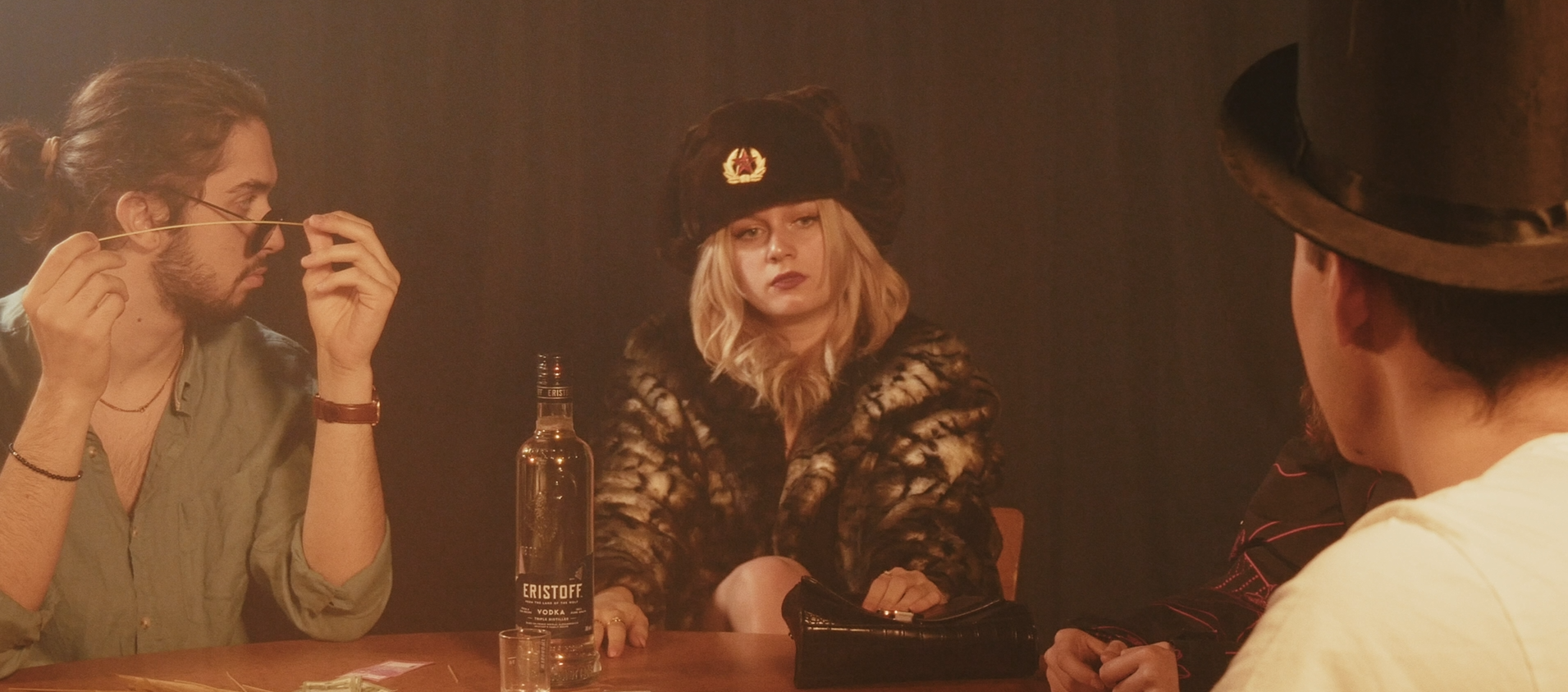 Scène avec plusieurs personnes assises autour d'une table. Une femme blonde au centre porte un chapeau militaire noir et un manteau de fourrure, une bouteille de vodka est posée sur la table.