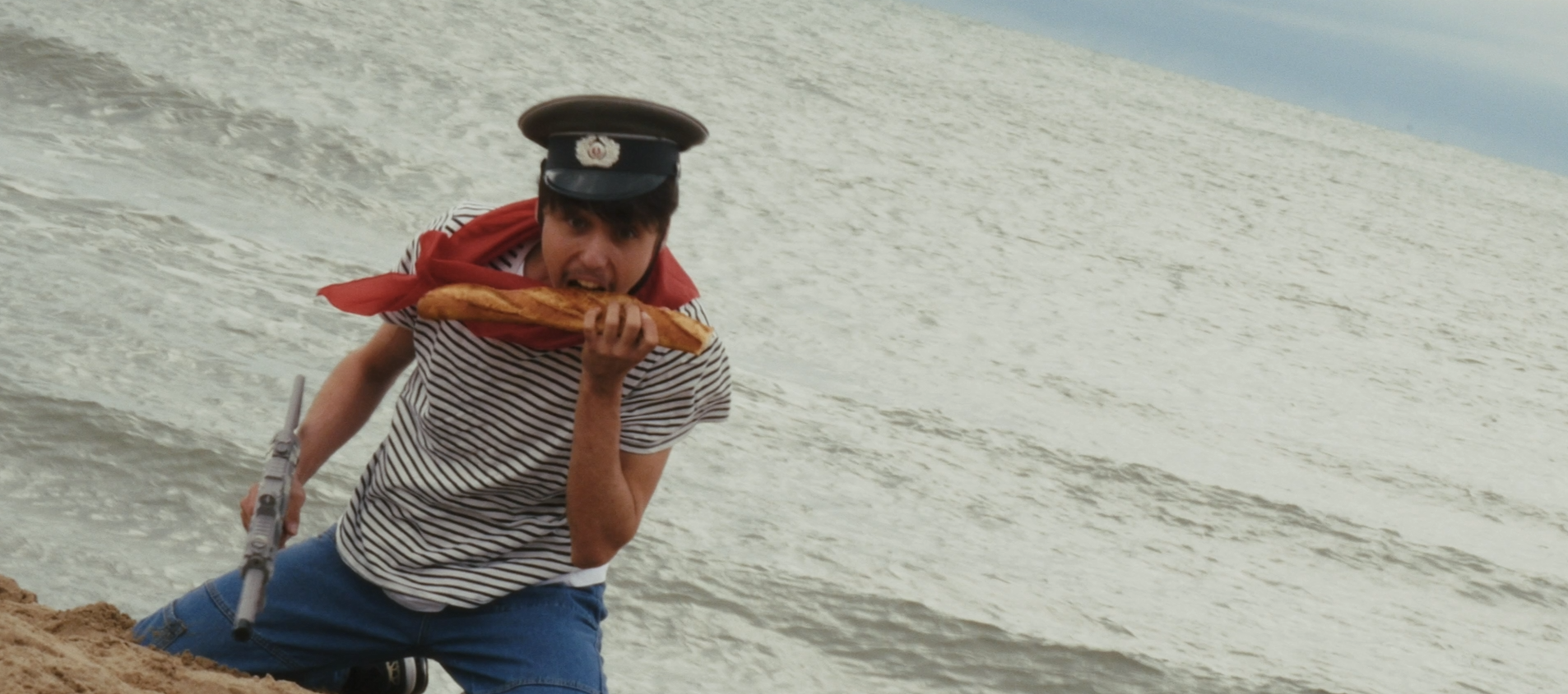 Homme portant une marinière et un chapeau militaire, tenant une baguette dans sa bouche et un fusil jouet à la main, sur une plage.